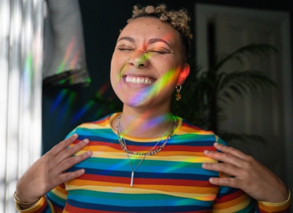 Young woman in rainbow top smiles at rainbow light in her window